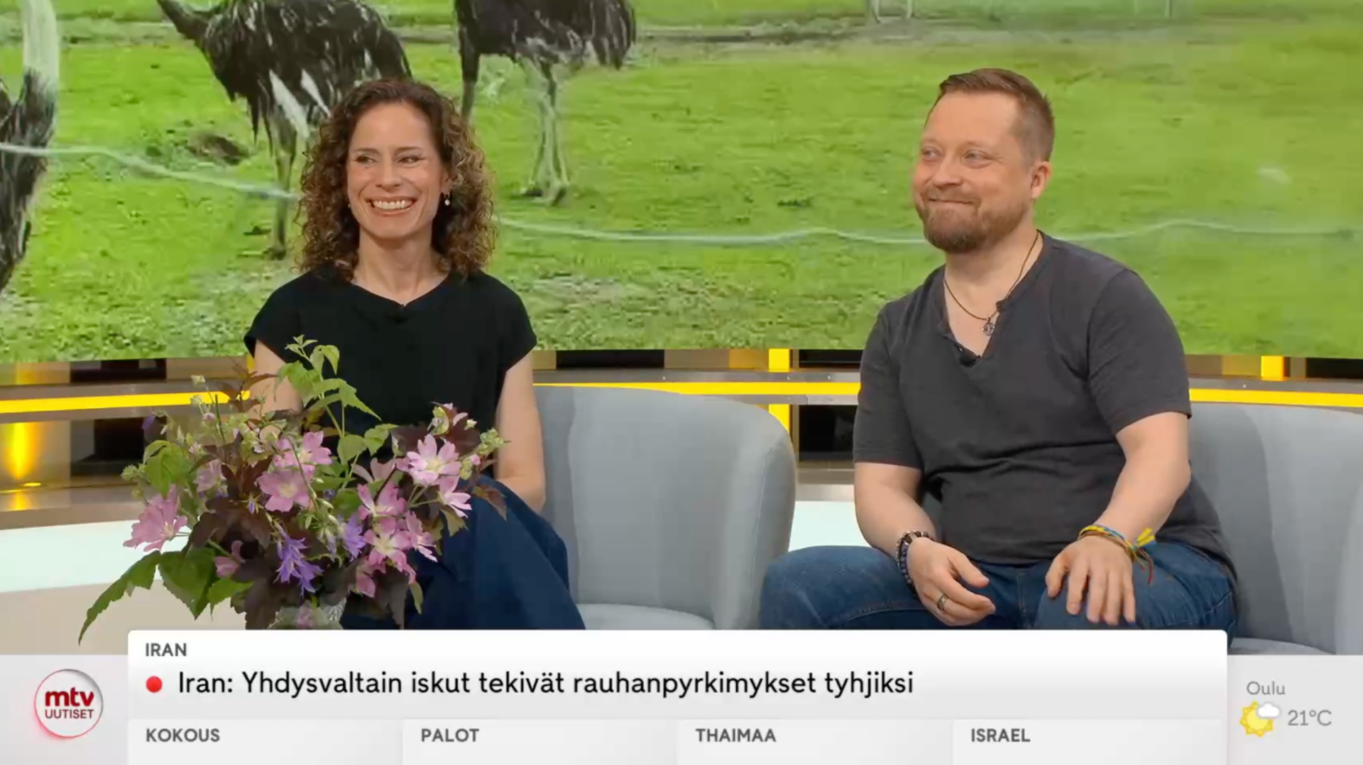 Two people sit on a studio couch outdoors, smiling, with a bouquet of flowers in foreground.